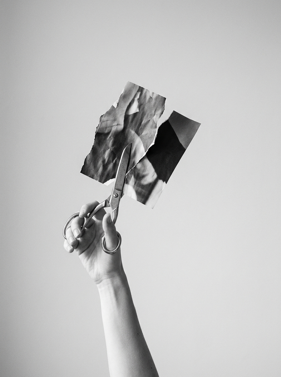 A hand raised against a grey background, cutting a torn photograph with scissors